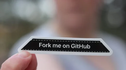 Close-up of a hand holding a 'Fork me on GitHub' sticker, blurred background.
