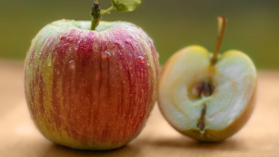 Close-up of a fresh red apple with dew, beside a sliced half on a wooden surface.