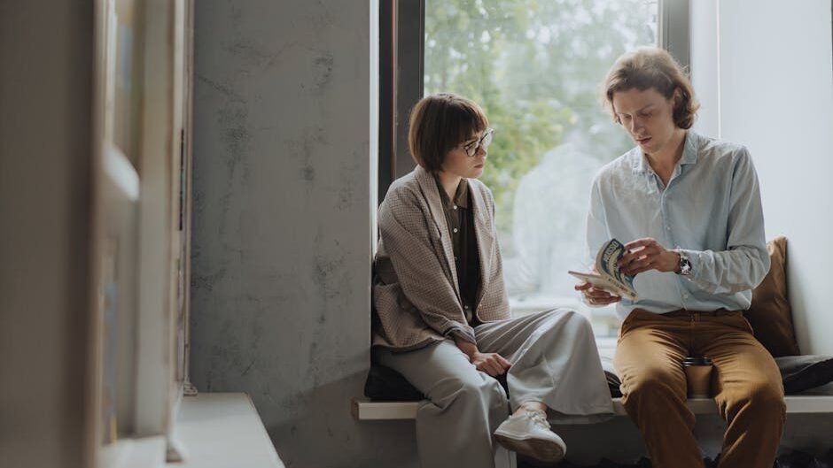 Two young adults enjoy a book in a cozy window seat, ideal for intellectual leisure.