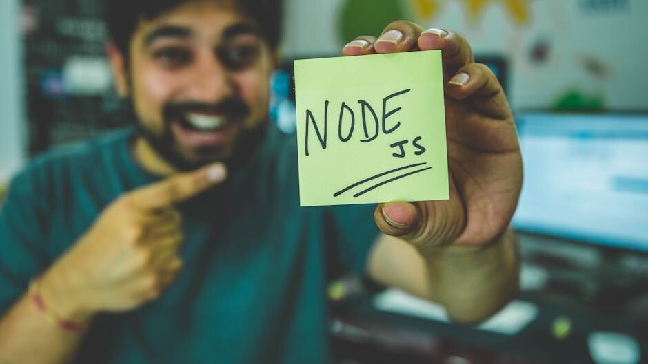 A cheerful software developer shows a Node.js sticky note in a tech workspace.