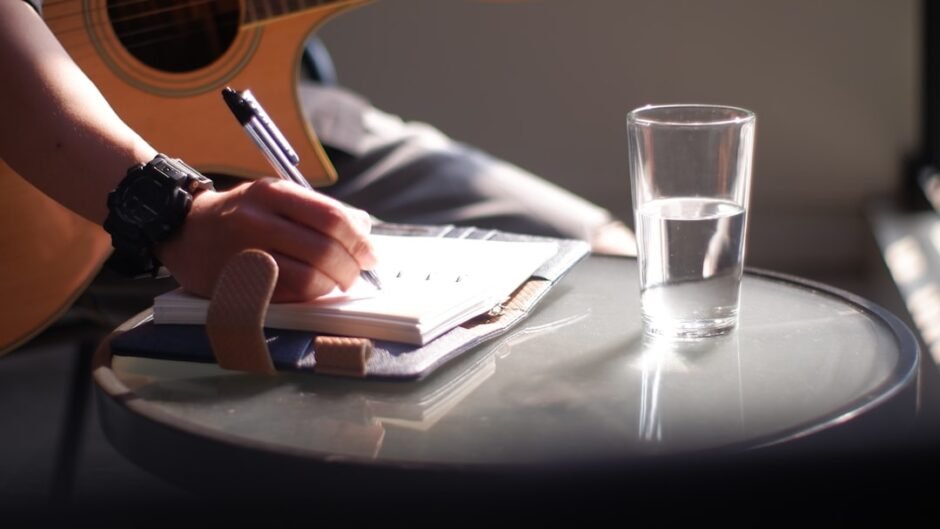 person holding pen writing on white paper beside clear drinking glass